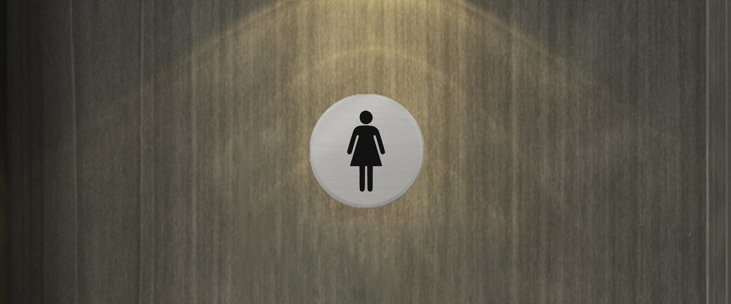 Rundes WC Schild in gebürsteter Silber-Optik mit schwarzem Damen-Symbol an einer dunklen Holztür im warmen Licht