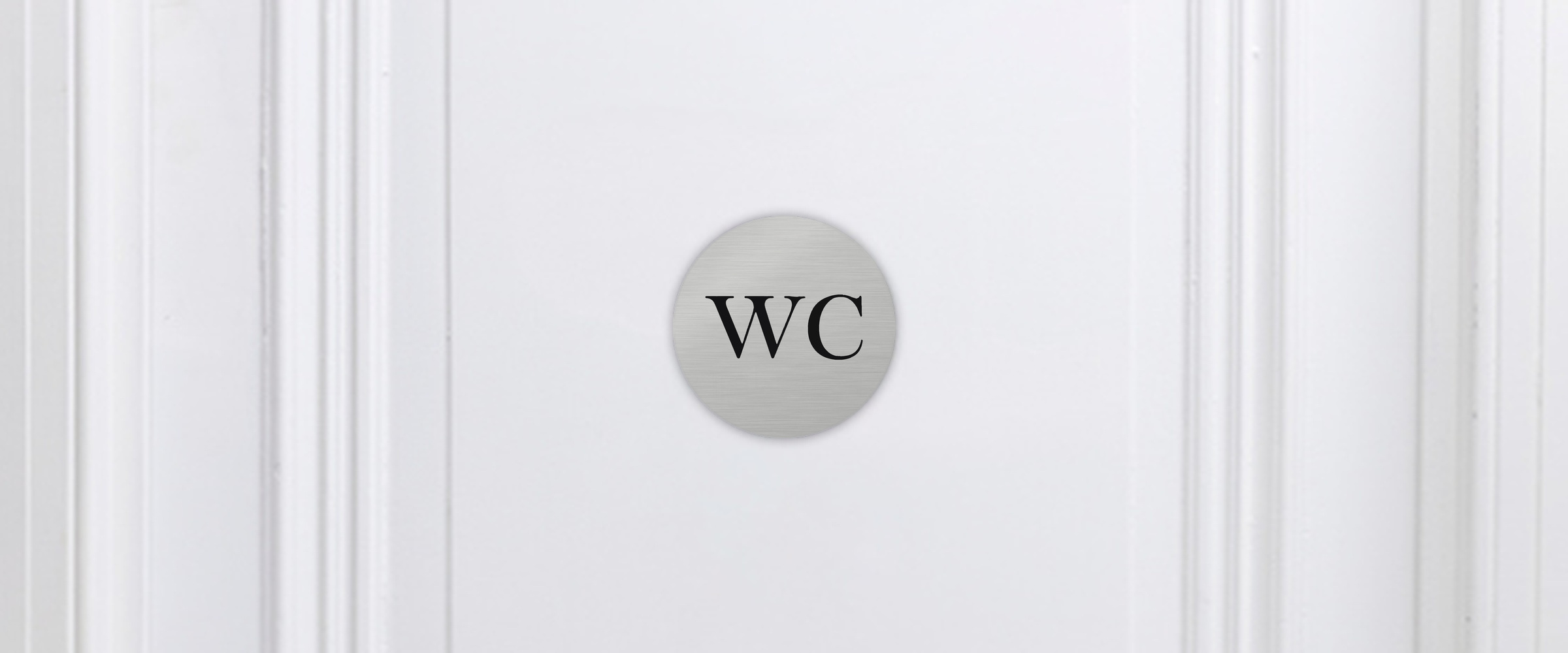 Rundes WC Schild in gebürsteter Silber-Optik mit schwarzem Schriftzug „WC“ an einer weißen Kassettentür