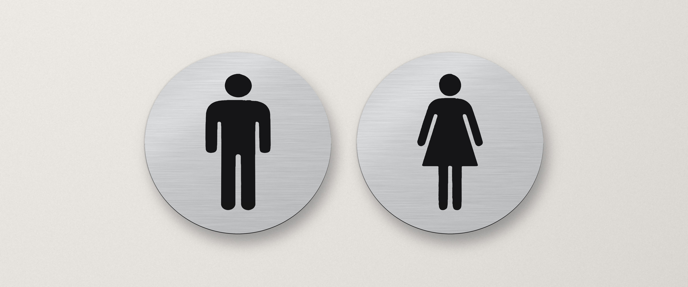 Zwei runde WC-Schilder in gebürsteter Silber-Optik an einer hellen Wand: Herren-Symbol und Damen-Symbol in Schwarz