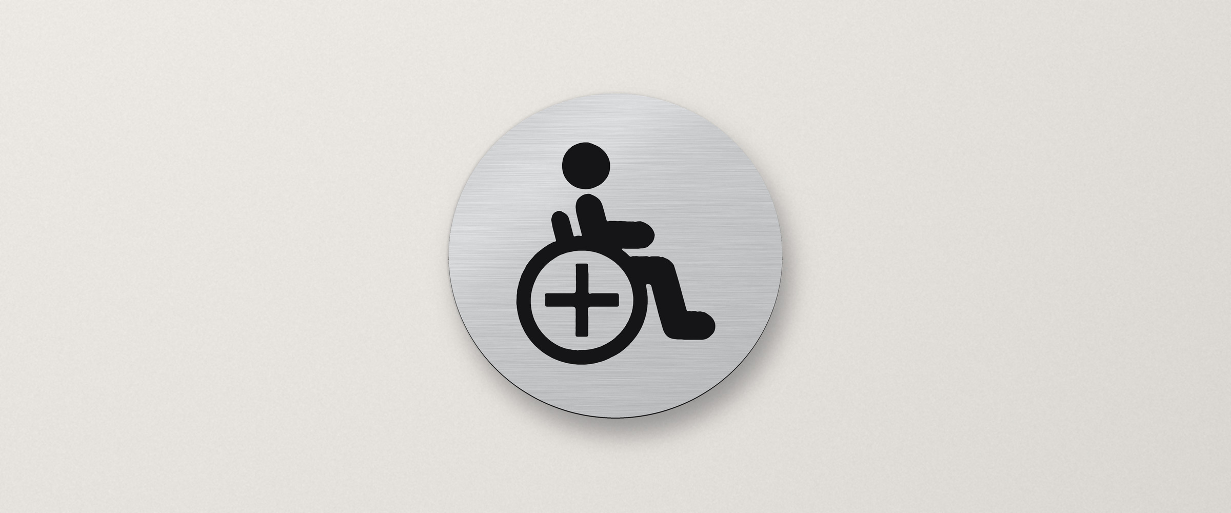 Rundes Schild in gebürsteter Silber-Optik an einer hellen Wand mit schwarzem Rollstuhl-Symbol für barrierefreie Toiletten