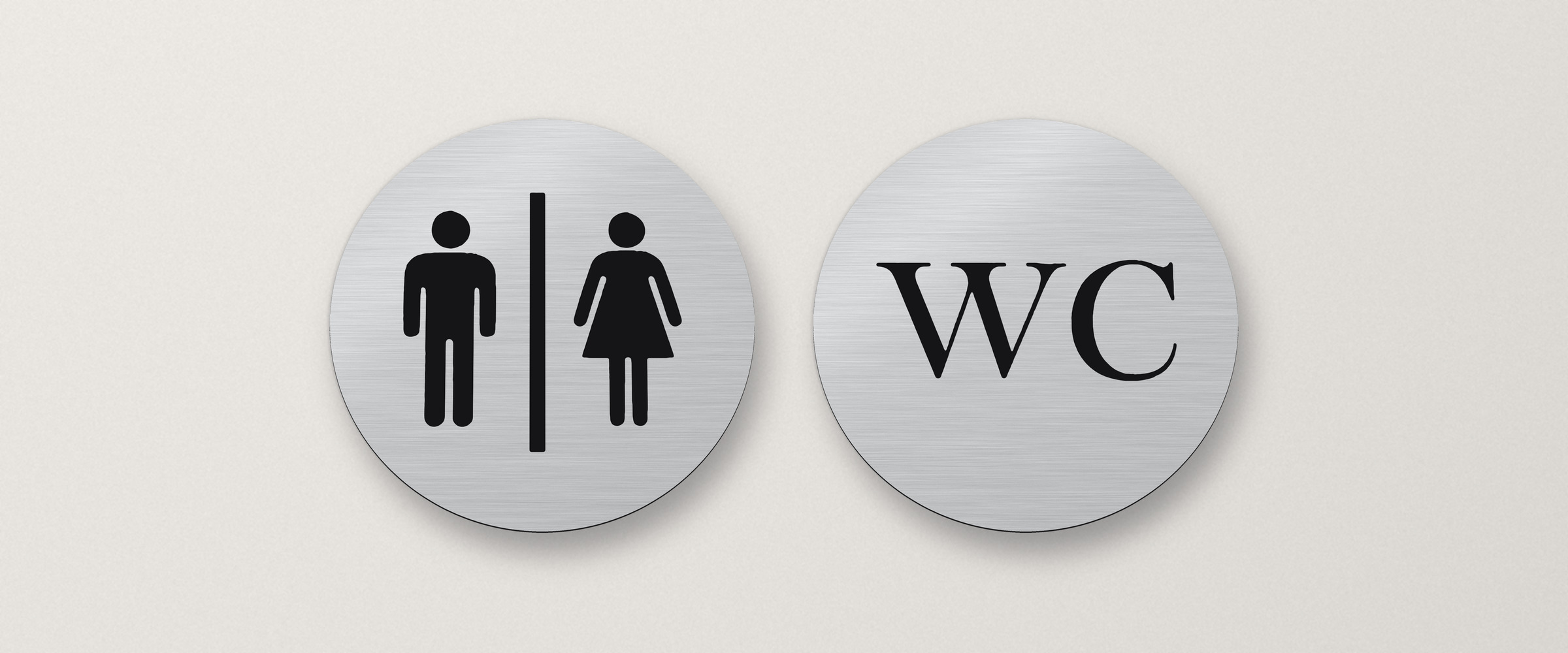 Zwei runde WC-Schilder in gebürsteter Silber-Optik an einer hellen Wand: Herren-und-Damen-Symbol und Schriftzug „WC“ in Schwarz